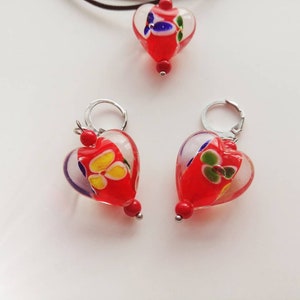 Puede incluir: Un conjunto de dijes de coraz&oacute;n de vidrio rojo con dise&ntilde;os florales blancos y coloridos. Los dijes est&aacute;n unidos a aros y una cadena de plata.