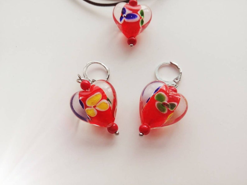 Puede incluir: Un conjunto de dijes de coraz&oacute;n de vidrio rojo con dise&ntilde;os florales blancos y coloridos. Los dijes est&aacute;n unidos a aros y una cadena de plata.