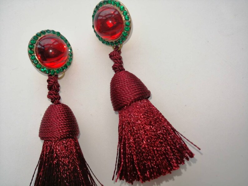 May include: A pair of red tassel earrings with green rhinestone accents. The earrings feature a round red stone surrounded by green rhinestones.
