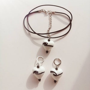 Puede incluir: Un conjunto de pendientes de vidrio en forma de coraz&oacute;n blanco y un collar con dise&ntilde;os de espiral negros. El collar est&aacute; en un cord&oacute;n negro con una cadena plateada.