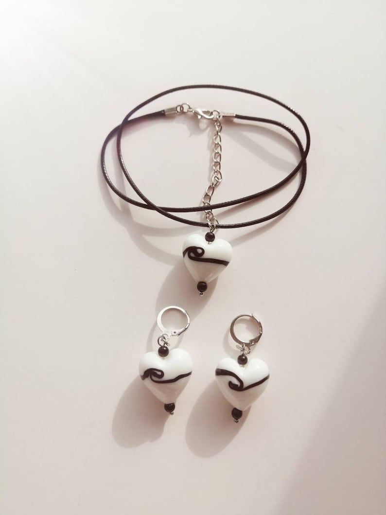 Puede incluir: Un conjunto de pendientes de vidrio en forma de coraz&oacute;n blanco y un collar con dise&ntilde;os de espiral negros. El collar est&aacute; en un cord&oacute;n negro con una cadena plateada.