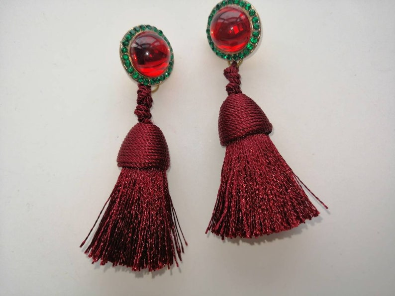 May include: A pair of red tassel earrings with green jeweled tops. The tassels are made of a soft, silky material.