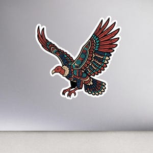 May include: A colorful sticker featuring a stylized eagle with outstretched wings. The eagle has a red head, brown and teal wings, and intricate patterns. The sticker is on a laptop, with a mouse, notebook, pencil, glasses, and small plants nearby.