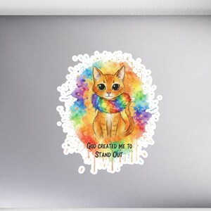 Puede incluir: Una pegatina con una ilustración de acuarela de un gato naranja con una bufanda arcoíris. El gato está centrado en un fondo blanco con salpicaduras de pintura coloridas. El texto "GOD CREATED ME TO STAND OUT" está debajo.