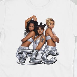 May include: A white short-sleeved t-shirt featuring a graphic print of the musical group TLC. The design shows three individuals in silver outfits positioned above the metallic, chrome-styled text reading TLC in a bold, rounded font.