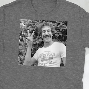 Puede incluir: Una camiseta gris jaspeado con una imagen en blanco y negro de un hombre haciendo el signo de la paz. El hombre lleva una camiseta blanca con texto. Una cámara vintage y otros accesorios están en el fondo.