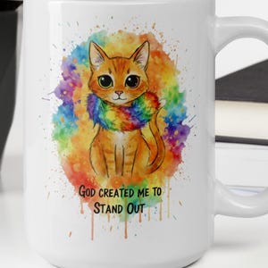 Puede incluir: Taza de cerámica blanca con una ilustración de acuarela de un gato naranja con un collar de arcoíris. La taza tiene el texto "GOD CREATED ME TO STAND OUT". El fondo tiene salpicaduras de pintura coloridas.