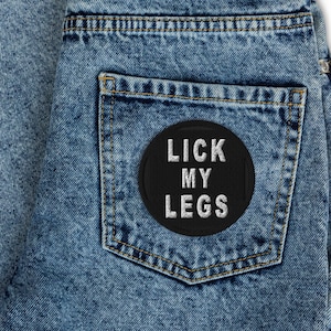 May include: A pair of blue denim jeans with a black patch on the back pocket that says "Lick My Legs" in white lettering.