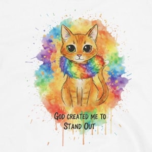 Puede incluir: Camiseta blanca con una ilustración de acuarela de un gato naranja con una bufanda de arcoíris. El gato está centrado contra un fondo de salpicaduras de colores. El texto "GOD CREATED ME TO STAND OUT" está impreso debajo.