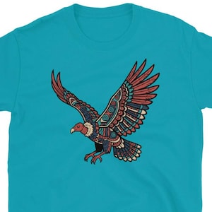 May include: A turquoise t-shirt featuring a detailed illustration of a soaring eagle. The eagle is depicted with outstretched wings, showcasing a vibrant design in shades of red, blue, and beige. The bird's head is red, and the design has a tribal art style.