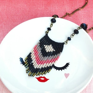 Op de afbeelding: Een zwarte, roze en witte kralenketting met een franje hanger hangt aan een wit keramiek schaaltje met een gestileerd gezicht ontwerp. De ketting is aan een gouden ketting.