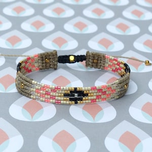 Puede incluir: Una pulsera tejida con un patrón geométrico. La pulsera presenta una base beige con filas de cuentas rosas, negras y doradas. Es ajustable con cordones beige y detalles dorados.