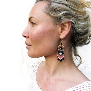 May include: A woman with blonde hair wearing a pair of black and coral beaded earrings with a black bead and gold circle accent.