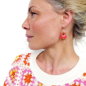 May include: A woman wearing a white crocheted sweater with a colourful granny square pattern. The sweater has a pink, orange and yellow colour scheme. She is wearing pink and gold dangle earrings.