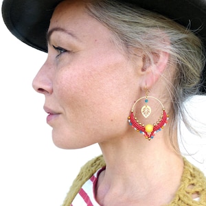 Op de afbeelding: Een vrouw draagt een rood-wit gestreept shirt en een bruine hoed met een paar rood-gouden oorringen met een bladerenontwerp en kleurrijke kralen.