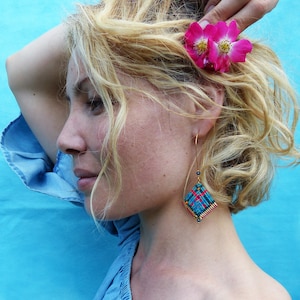 Peut inclure: Une femme aux cheveux blonds porte une fleur rose dans les cheveux, un haut bleu à épaules dénudées et des boucles d'oreilles en macramé colorées. Les boucles d'oreilles sont en forme de losange avec des fils bleus, rouges et oranges.