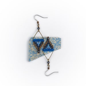 以下が含まれることがあります： 幾何学模様の青いビーズと黒いビーズのイヤリングのペア。イヤリングはシルバーのフックにぶら下がっています。