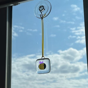 May include: A glass bee suncatcher with a yellow and black striped body and a purple bow hanging from a window with a suction cup.