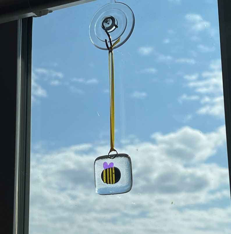 May include: A glass bee suncatcher with a yellow and black striped body and a purple bow hanging from a window with a suction cup.
