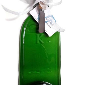 May include: A green glass bottle shaped like a serving tray with a cork and a small knife. The bottle is engraved with the letter "K".