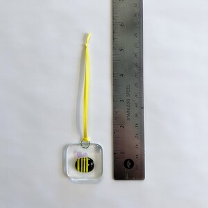 May include: A small glass ornament with a yellow and black bee design. The ornament is attached to a yellow ribbon.