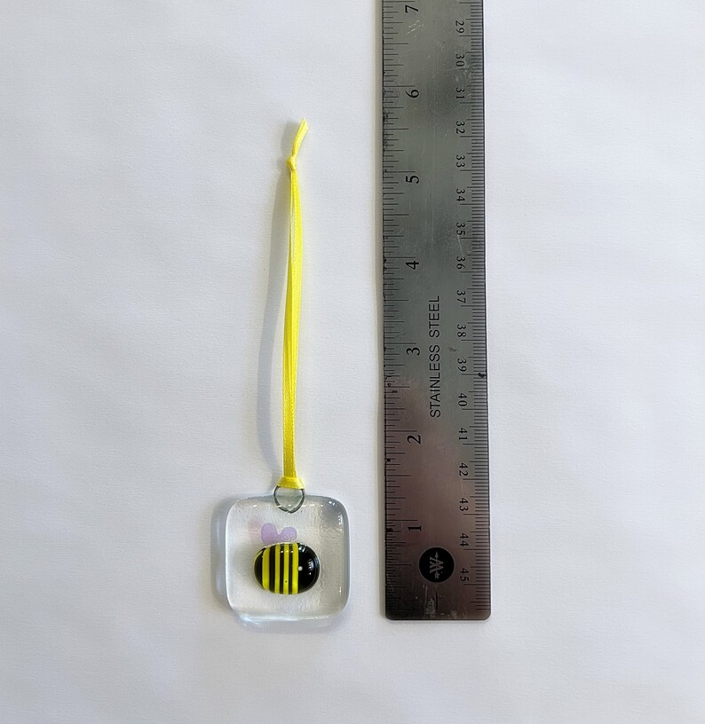 May include: A small glass ornament with a yellow and black bee design. The ornament is attached to a yellow ribbon.