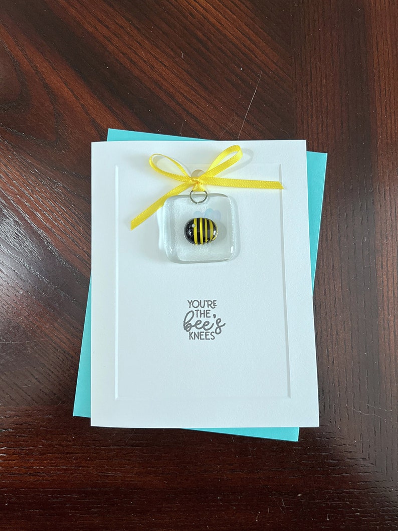 May include: A white card with a yellow ribbon tied around a small glass square with a bee inside. The card says "You're the bee's knees".