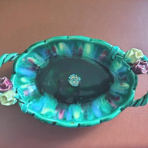 May include: Oval ceramic serving dish with a glossy, mottled green and dark interior. The dish has two twisted rope handles and is adorned with rose-shaped accents in yellow and burgundy. A small emblem is visible in the centre.