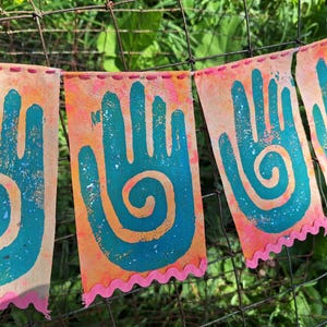 May include: A decorative bunting banner featuring four rectangular fabric flags. Each flag has a peach and orange mottled background with a teal handprint graphic containing a central spiral. The bottom edges feature pink rickrack trim.