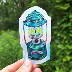 May include: A teal and green sticker of a vintage Coleman lantern. The lantern has a detailed design with a teal top, a glass enclosure, and a green base. The sticker has a holographic effect and the text "Coleman" is visible.