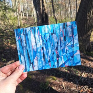 May include: A rectangular watercolor painting with an abstract design. The artwork features a mix of blue shades, with black and pink accents. The painting is held in front of a blurred forest background.