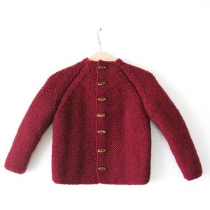 May include: A deep red, long-sleeved knit cardigan featuring a textured garter stitch pattern. The garment is fastened down the center front with six brown, oblong wooden toggle buttons and is displayed on a light-colored wooden hanger.