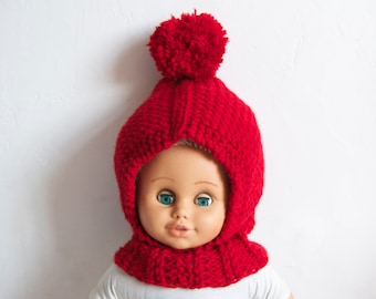 May include: A red knitted hat with a pom-pom on top. The hat has a fold-down flap that covers the neck and chin. The hat is worn by a doll with blue eyes.