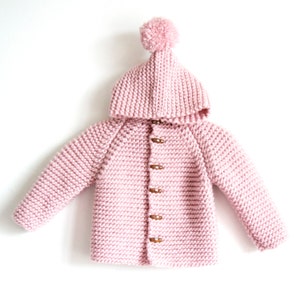 May include: A light pink, long-sleeved, hooded knit cardigan featuring a textured garter stitch pattern. The garment includes a matching pom-pom on the top of the hood and is fastened down the center front with five brown, oval-shaped wooden toggle buttons.
