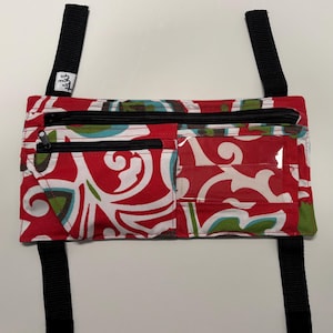 May include: A red and white patterned fanny pack with black zippers and straps. The pack has two zippered compartments and a clear pocket. The straps are black and have a small white tag with black text.