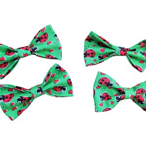 May include: Four green bow ties featuring a pattern of red ladybirds and red hearts. The bow ties are displayed against a white backdrop. The ladybirds have black spots and antennae.