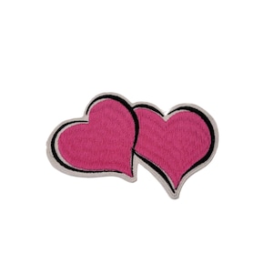 Puede incluir: Dos corazones rosas con contornos negros, cosidos juntos, son un diseño lindo y simple para un parche.