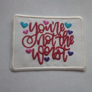 May include: Embroidered patch with the phrase "you're not the worst" in red script. The white patch has a cream-colored border and small heart accents in pink, teal, and purple.