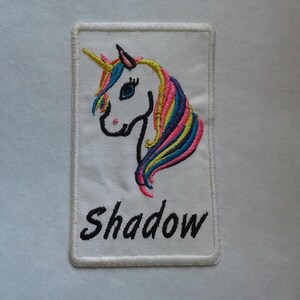 Puede incluir: Parche blanco bordado con una cabeza de unicornio arcoíris y la palabra "Shadow" en letras negras.