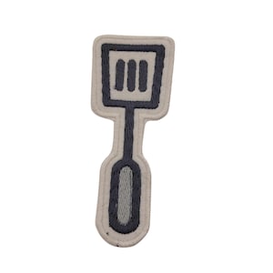 Peut inclure: Écusson brodé représentant une spatule. Le patch a un fond gris clair avec un contour et une poignée de spatule gris foncé. La tête de la spatule comporte trois fentes verticales. Le patch est bordé de gris clair.