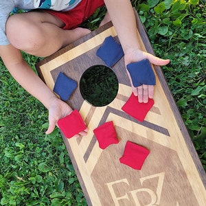 Op de afbeelding: Een houten cornhole-spelbord met een zwart gat en een rood-wit-blauw ontwerp. Op het bord staat de tekst "EBZ Cornhole LLC". Een kind speelt het spel met rode en blauwe bonenzakken.
