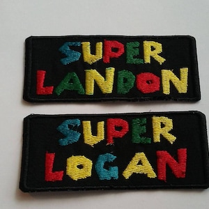 Pode incluir: Dois remendos de tecido preto com costuras brancas. O remendo superior diz "SUPER LANDON" em letras coloridas. O remendo inferior diz "SUPER LOGAN" em letras coloridas.