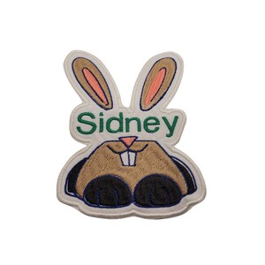 May include: Embroidered patch in the shape of a bunny rabbit with the name "Sidney" in green lettering. The bunny has brown fur, pink inner ears, and black paws. The patch is outlined in white and blue.