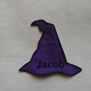 Könnte beinhalten: Lilafarbener Filzhut mit schwarzer Naht und dem Namen "Jacob" auf der Vorderseite gestickt.
