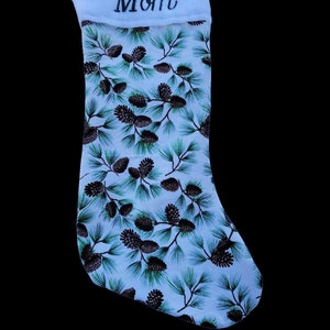 Puede incluir: Un calcetín navideño con un puño blanco bordado con la palabra "Mamá". El cuerpo del calcetín presenta un fondo azul claro con un estampado de ramas de pino y piñas.