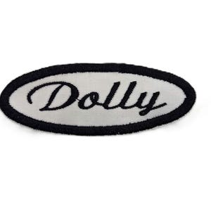 Puede incluir: Un parche bordado de forma ovalada con fondo blanco y un borde negro grueso. La palabra Dolly está bordada en el centro con una fuente cursiva negra. El parche tiene una superficie texturizada y bordes definidos en todo el diseño.
