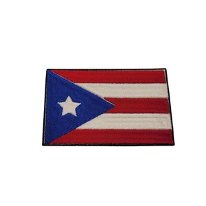 Può includere: Una toppa ricamata con la bandiera di Porto Rico. La bandiera è rossa, bianca e blu con una stella bianca su un triangolo blu nell'angolo in alto a sinistra.