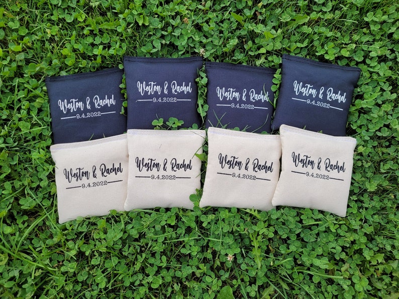 May include: Eight cornhole bags, four navy and four cream, are displayed on green grass. Each bag features the names "Weston & Rachel" and the date "9.4.2022" printed in a cursive style, suitable for a wedding or event.