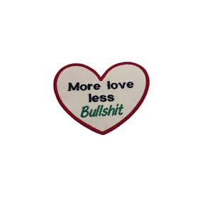 May include: Heart-shaped patch with a white center, a red border, and the text "More love less Bullshit" in navy blue and green. The patch is likely for clothing or accessories.