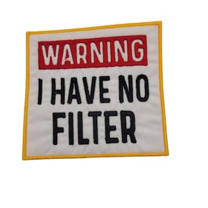 May include: A white embroidered patch with a yellow border. The patch has red text that says "WARNING" and black text that says "I HAVE NO FILTER".